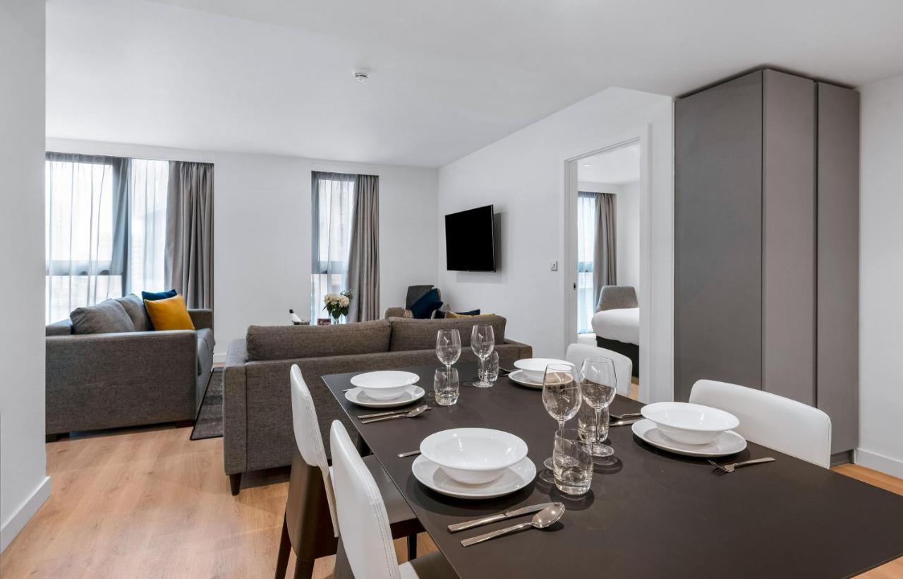 Citysuites 2 Aparthotel 曼彻斯特 外观 照片 The photo shows a modern living space with an open layout. In the foreground, there is a dining table set for a meal, featuring multiple plates, glasses, and cutlery arranged neatly. In the background, there is a comfortable-looking gray sofa with a 