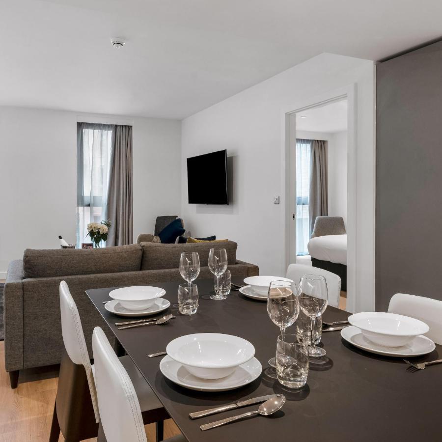 Citysuites 2 Aparthotel 索尔福德 外观 照片 The photo shows a modern, well-lit living space that features a dining area and a sitting area. In the foreground, there is a dining table set for six, with white plates and crystal glasses arranged neatly. The table is surrounded by white chairs. 

