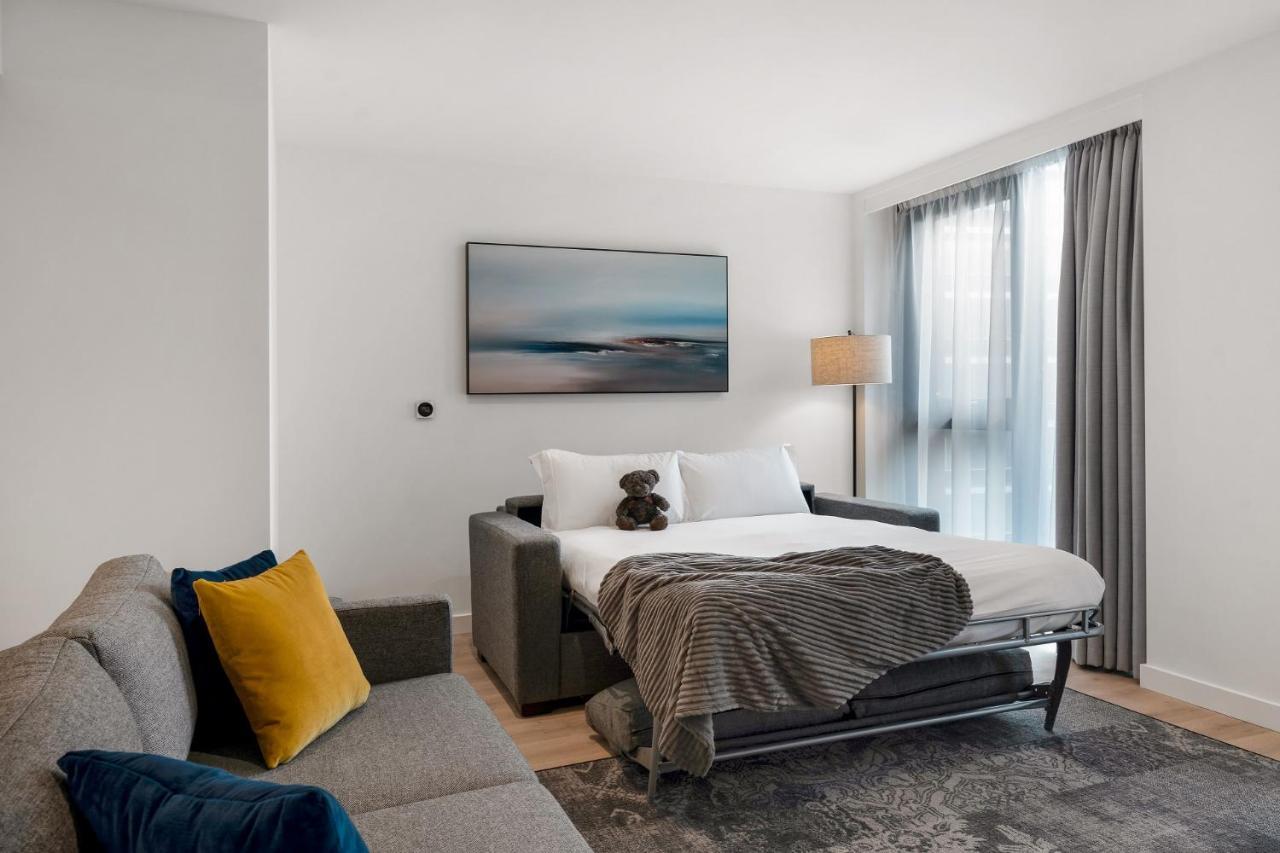 Citysuites 2 Aparthotel 索尔福德 外观 照片 The photo shows a modern and minimalistic guest room. There is a sofa on the left side, adorned with a colorful cushion. In the middle is a bed that appears to be a pull-out style, featuring white bedding and a gray throw blanket. A stuffed toy is pl