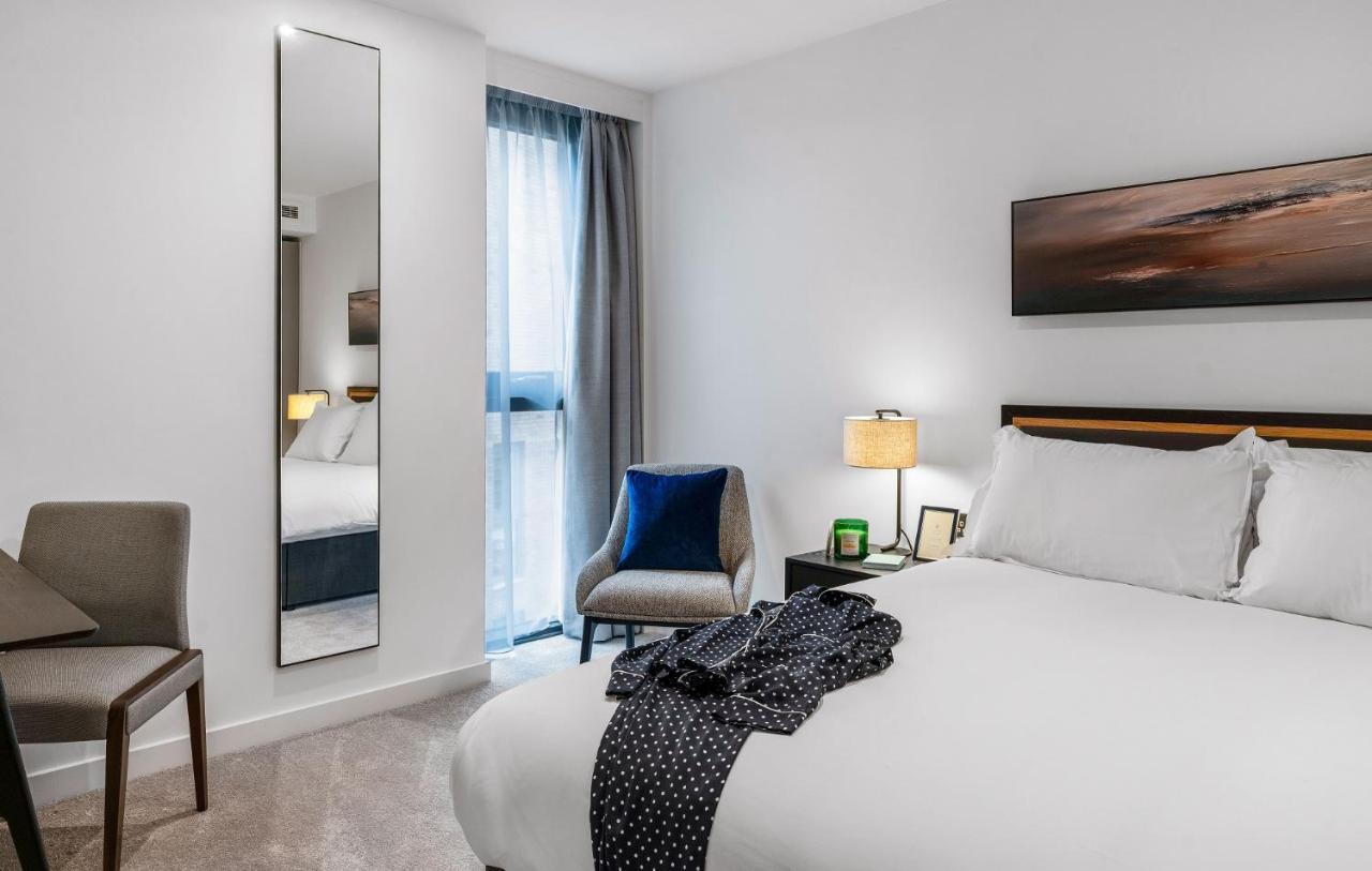 Citysuites 2 Aparthotel 曼彻斯特 外观 照片 The photo shows a modern hotel room. It features a neatly made bed with white linens and a dark-colored throw draped over it. There is a small bedside table with a lamp next to the bed. A comfortable chair in a light color is positioned near the wind