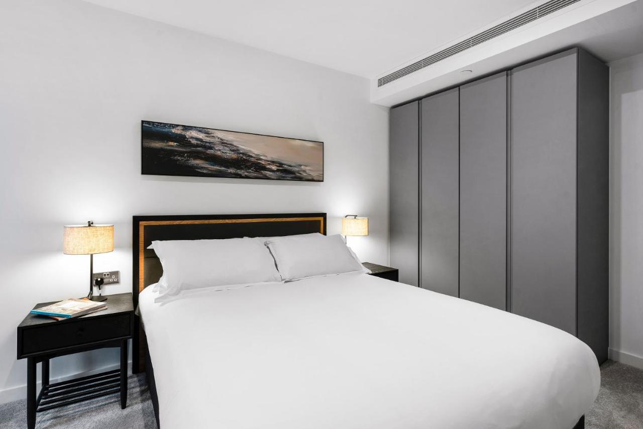 Citysuites 2 Aparthotel 曼彻斯特 外观 照片 The photo shows a modern bedroom designed with a minimalist aesthetic. The focal point is a large bed with white bedding, positioned against a wall. On either side of the bed, there are black nightstands topped with lamps, providing a warm glow. Abov