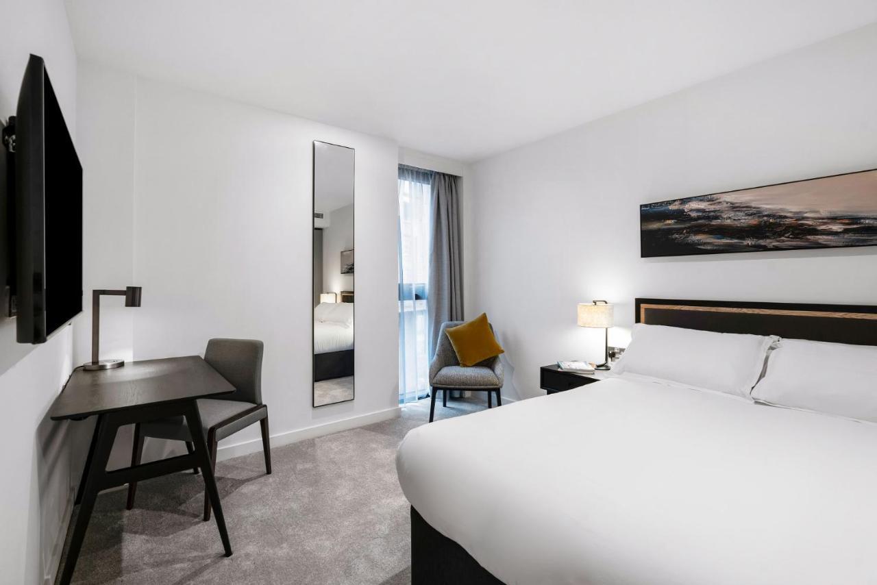 Citysuites 2 Aparthotel 曼彻斯特 外观 照片 The photo shows a modern hotel room with a clean and minimalist design. It features a large bed with white linens, a small desk with a chair, and a mirror mounted on the wall. There is a chair in one corner with a decorative pillow, and the room has 