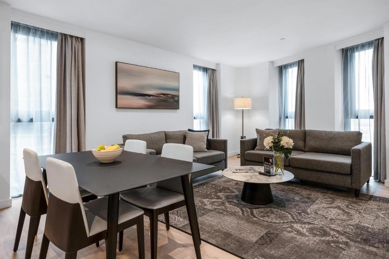 Citysuites 2 Aparthotel 曼彻斯特 外观 照片 The photo depicts a modern and stylish interior of a living space. On the left, there is a dining table with four chairs, featuring a bowl of fruits on the table. In the background, there's a cozy seating area with a gray sofa and a smaller round cof
