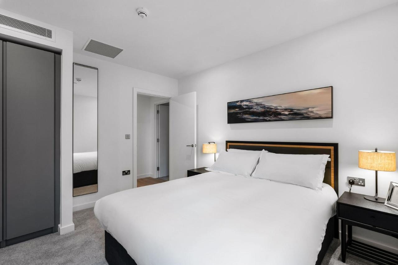 Citysuites 2 Aparthotel 曼彻斯特 外观 照片 The photo shows a modern bedroom featuring a large double bed with white bedding. There are two bedside tables with lamps on either side of the bed. The walls are painted white, and there's a piece of artwork above the bed that depicts a landscape. A