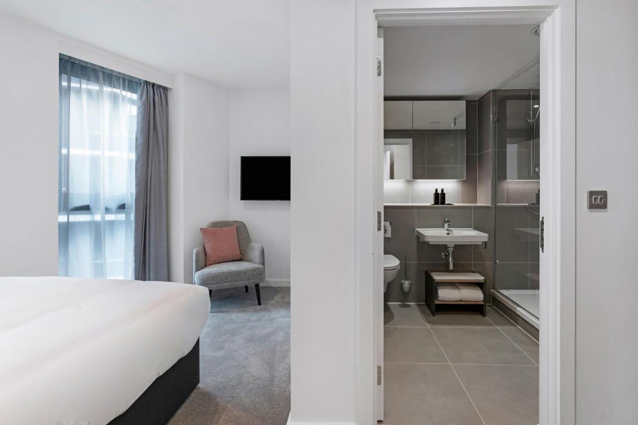Citysuites 2 Aparthotel 曼彻斯特 外观 照片 The photo shows a modern hotel room and bathroom setup. On the left, there is a bed with white bedding and a small sitting area that includes a gray chair and a pillow in a subtle pink shade. A flat-screen TV is mounted on the wall, providing enterta
