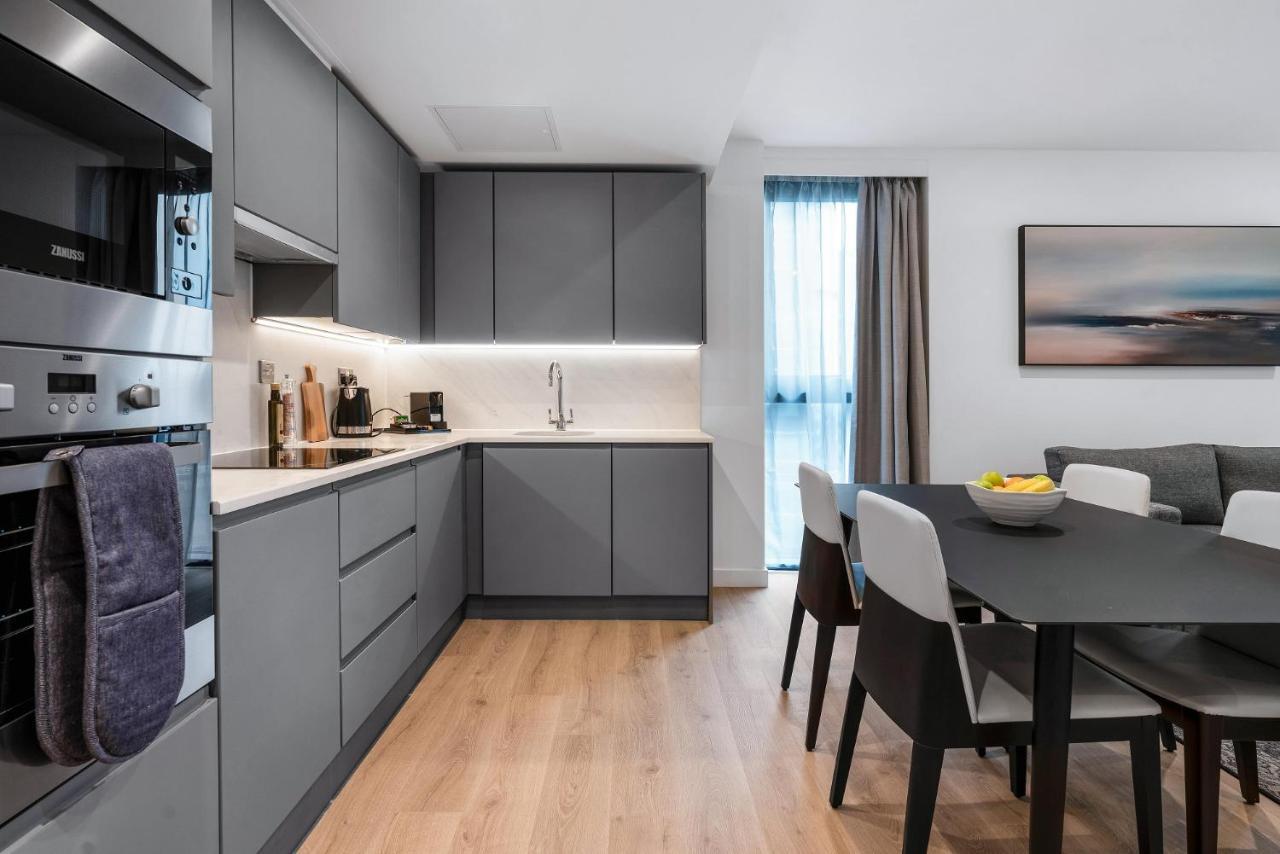 Citysuites 2 Aparthotel 曼彻斯特 外观 照片 The photo shows a modern kitchen and dining area. The kitchen features sleek gray cabinetry, a white countertop, and built-in appliances such as an oven. There is a sink and some kitchen utensils displayed. The adjacent dining area contains a black t