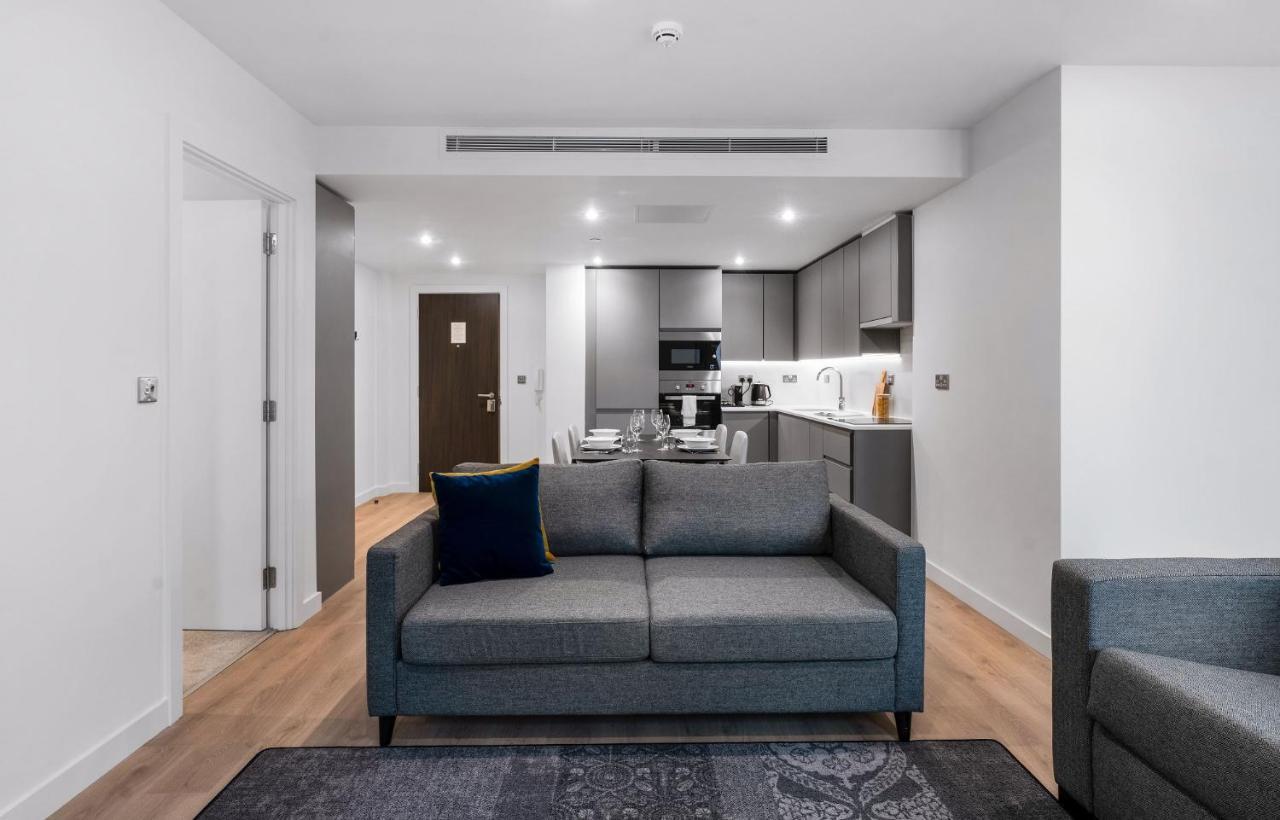Citysuites 2 Aparthotel 索尔福德 外观 照片 The photo shows a modern interior space, likely a living area or apartment. In the foreground, there is a gray sofa with a blue cushion positioned on a textured rug. To the back, there is an open kitchen with sleek cabinetry and appliances, visible a