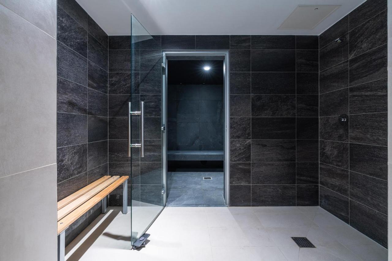 Citysuites 2 Aparthotel 索尔福德 外观 照片 The photo shows a modern, minimalist shower area. The space is predominantly tiled with dark gray tiles on the walls, giving it a sleek and sophisticated look. There is a glass door that leads to another room, and a wooden bench is positioned against