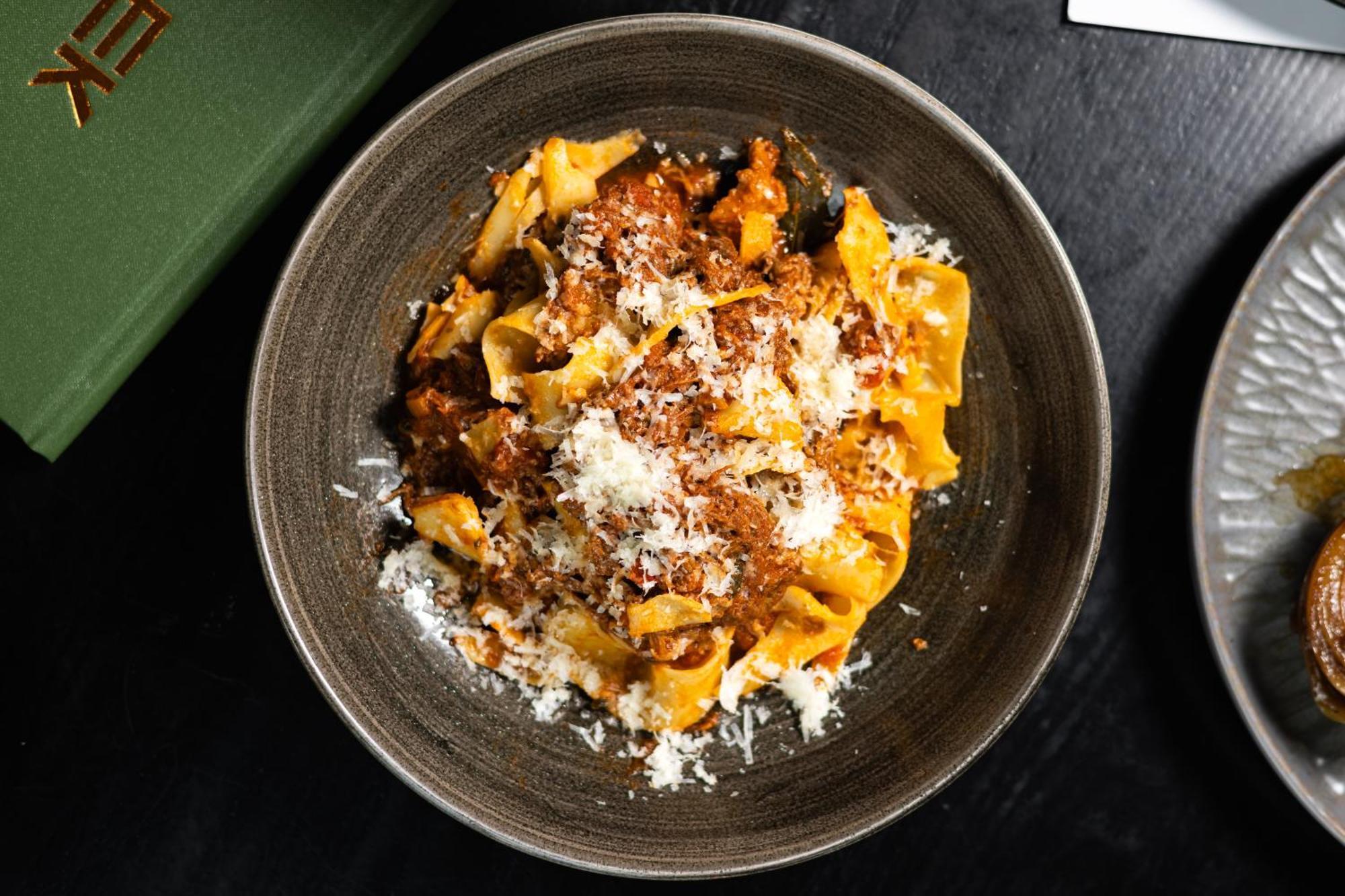 Citysuites 2 Aparthotel 索尔福德 外观 照片 The photo shows a bowl of pasta featuring wide, ribbon-like noodles, likely pappardelle, fully coated in a rich meat sauce. The dish is garnished with a generous sprinkling of grated cheese, adding a creamy texture to the presentation. The dark bowl 